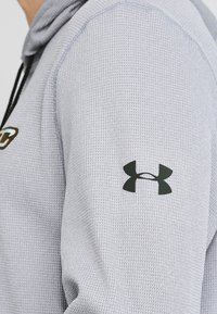 Primer plano de una persona que lleva una sudadera con capucha de manga larga texturada de color gris claro, con un logo negro de Under Armour en la parte superior del brazo.