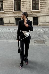 Črn blazer z belim prugastim pasom, črne hlače, ostri bordo petki in mala črna torbica; gladka tkanina in natančno zasnovan kroj.