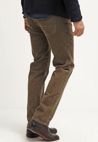 Mand iført brune fløjlsholdere, mørkeblå sweater og mørkebrune lædersko, stående mod en ensfarvet lys baggrund.