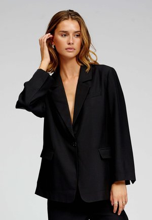 Czarna marynarka z klapami z wycięciem, zapięciem na jeden guzik, dwoma przednimi kieszeniami i luźnym krojem. Wykonana z gładkiej tkaniny o matowym wykończeniu.