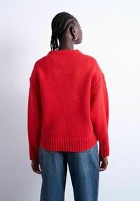 Personne aux cheveux tressés portant un pull en tricot rouge vif et un jean bleu, vue de dos sur un fond clair uni.