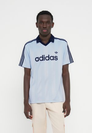 Camiseta Adidas azul claro con cuello azul marino y tres franjas en las mangas. Presenta un gran logo azul oscuro en el frente. Mangas cortas.