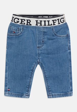 Short en denim bleu avec taille élastique, avec un texte noir en gras et un petit logo rouge, blanc et bleu sur la poche avant et l’ourlet.