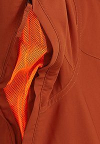 Bruine jasstof met een ritszak; aan de binnenzijde is helder oranje gaasmateriaal zichtbaar. Duidelijke stikseldetails op het kledingstuk.