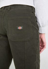 Tumši zaļi kordūra bikses ar aizmugurējo kabatu, kurai ir daudzkrāsains Dickies logo uzšuvums. Redzamā ribota tekstūra un šuvju detaļas.
