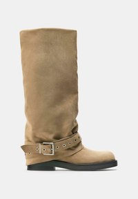Botte mi-haute en daim couleur chamois avec bout rond, petit talon noir et sangle décorative avec boucle argentée et œillets autour de la cheville.