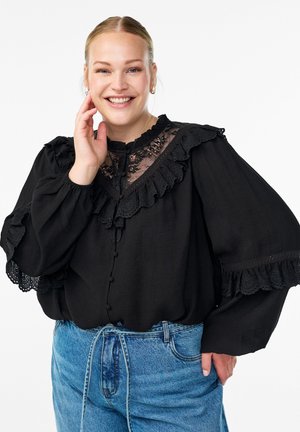 Smiling woman with blonde hair wearing a black lace ruffled blouse and light blue high-waisted jeans, posing with hand near face.