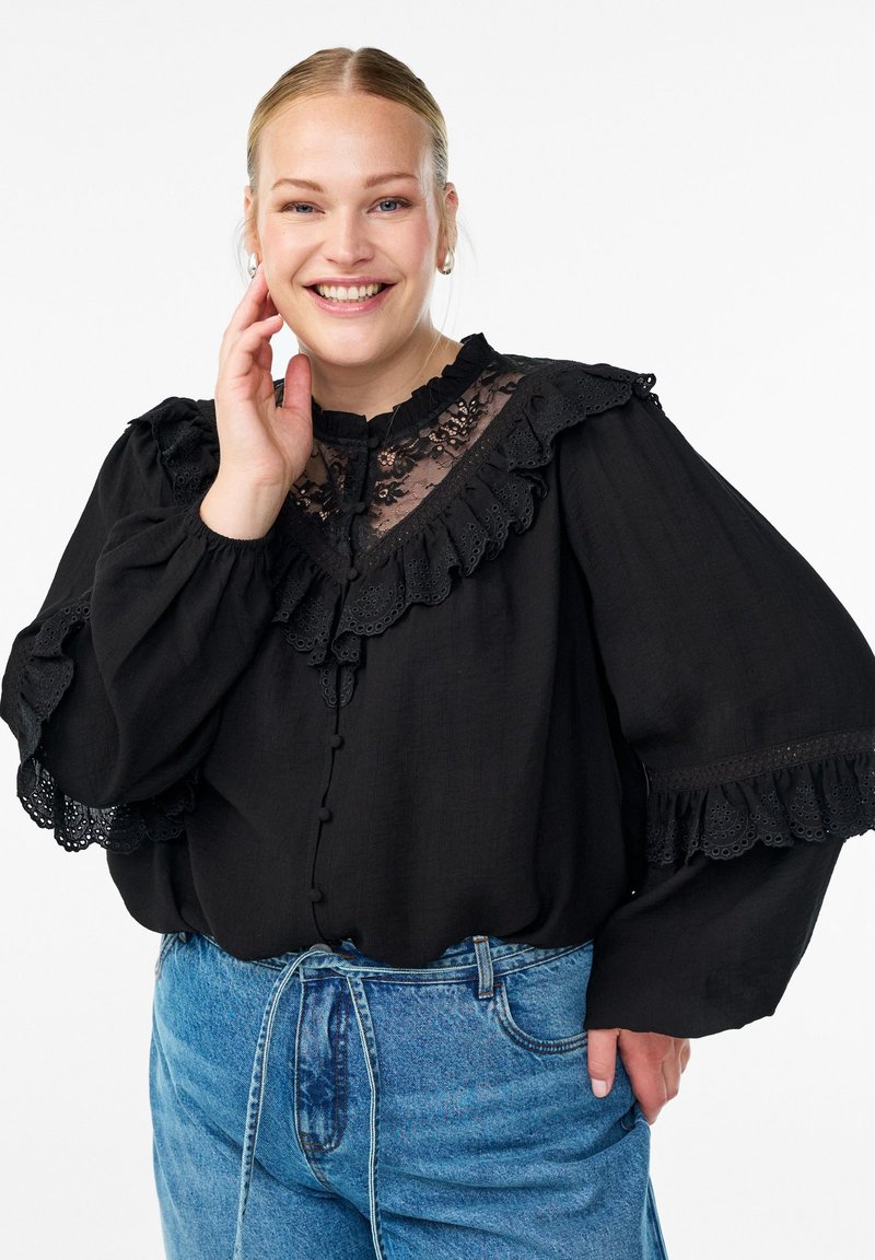 Lächelnde Frau mit blonden Haaren, die eine schwarze Spitzenbluse mit Rüschen und hellblaue hochtaillierte Jeans trägt, posiert mit der Hand nahe dem Gesicht.