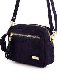 Námořnické modré semišové crossbody taška se zlatým kováním, obsahující hlavní zipovou kapsu, přední zipovou kapsu a dekorativní detail s třásněmi.