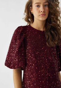 Top burgundy cu paiete, cu manșete scurte tip bufant, având o suprafață texturată cu paiete rotunde strălucitoare și un guler clasic rotund.