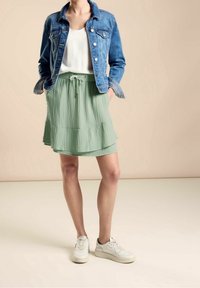 Džínová bunda, bílý top bez rukávů a vrstvená zelená sukně se strukturálními detaily. Obsahuje stahovací pas a bílé tenisky.