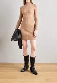 Robe en tricot beige côtelé avec col haut, manches longues et silhouette ajustée. Une veste en cuir noire et des bottes de cowboy complètent l'ensemble.