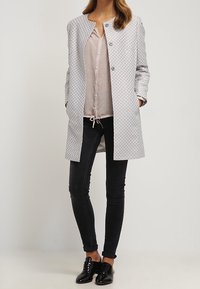 Femme portant un manteau long à motifs, un chemisier rose clair boutonné, un jean skinny noir et des chaussures Oxford noires, debout les mains dans les poches.