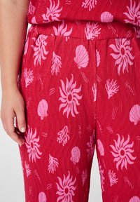 Pantalon rouge avec une texture froissée, arborant un motif de corail rose et de coquillages. Taille élastique et main reposant légèrement sur le tissu.