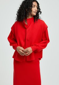 Rote gestrickte Jacke mit Ballonärmeln und Knopfverschluss, kombiniert mit einem passenden roten Oberteil und Rock, alles in einem glatten, strukturierten Stoff.