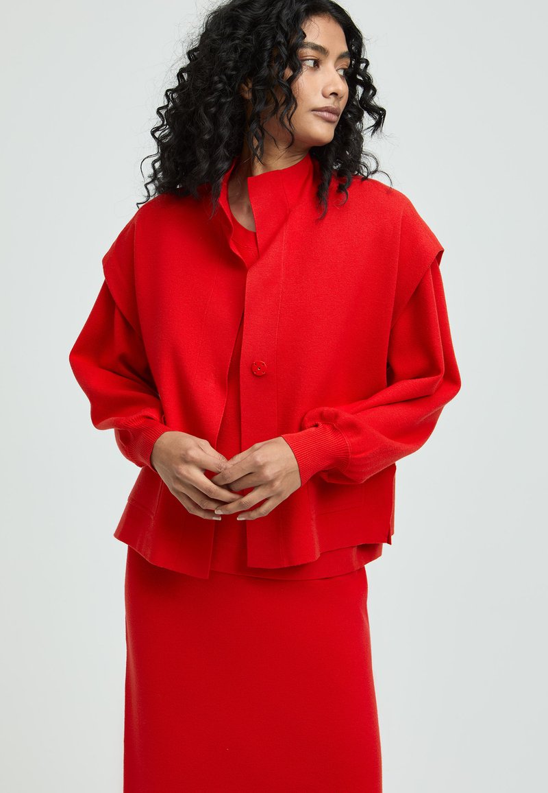 Rote gestrickte Jacke mit Ballonärmeln und Knopfverschluss, kombiniert mit einem passenden roten Oberteil und Rock, alles in einem glatten, strukturierten Stoff.