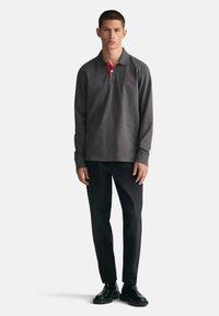 Langärmliges graues Poloshirt mit einem roten Kragen, kombiniert mit schwarzen Jeans und schwarzen Schuhen. Das Shirt hat ein kleines Logo auf der Brust.