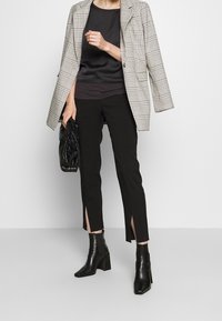Blazer im grauen Karomuster, schwarzes Satin-Oberteil, schwarze Hose mit seitlichen Schlitzen und schwarze, mit Absatz versehene Stiefeletten. Schwarze Clutch-Tasche in der Hand.
