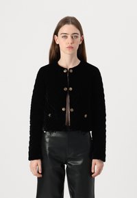 Giacca cropped in velluto nero con un design trapuntato, dotata di bottoni dorati e maniche lunghe. Abbinata a pantaloni in pelle nera.