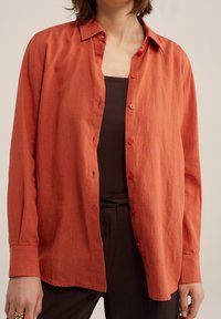 Rostfarbendes, knöpfbares Hemd mit lockerer Passform, aus leichtem Stoff. Mit Kragen, langen Ärmeln und roten Knöpfen.