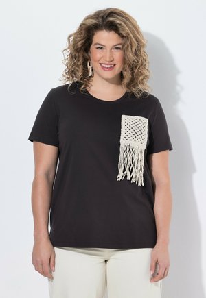 Frau mit lockigem Haar lächelt, trägt ein schwarzes Kurzarmshirt mit einer weißen Makramee-Tasche und hellfarbige Hosen, steht vor einer weißen Wand.