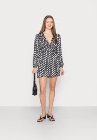 Vestido preto envolvente com padrões geométricos brancos, mangas longas e decote em V profundo, combinado com saltos pretos de tiras e uma bolsa pequena.