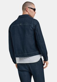 Giacca in denim scuro da uomo con taglio classico, dotata di colletto strutturato, polsini con bottone e un'etichetta discreta sul retro.