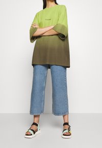 Olivově zelené a světle zelené gradientové oversized tričko, v kombinaci s modrými zkrácenými džíny a vícebarevnými sandály s masivní podrážkou.