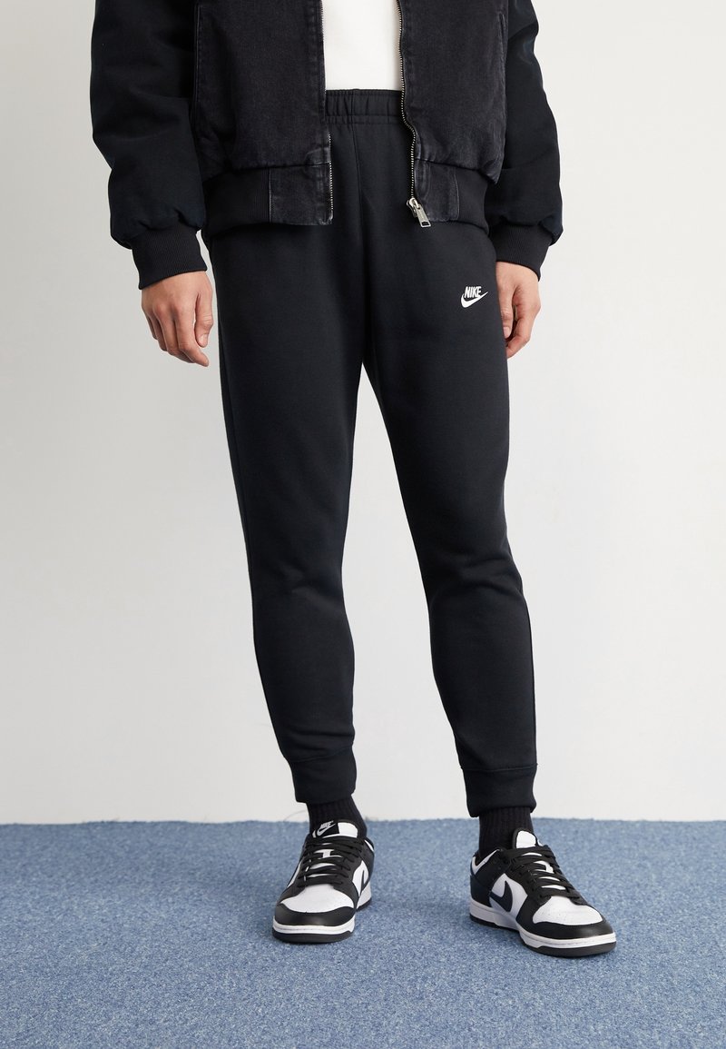 Pantalones de chándal negros de Nike con cintura elástica y logo, combinados con zapatillas en blanco y negro. El fondo presenta una superficie texturizada azul.