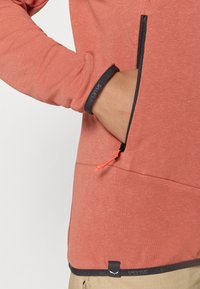 Μακρυμάνικο, κοραλλί πουλόβερ από μαλακό ύφασμα.  Διαθέτει τσέπη με φερμουάρ και πορτοκαλί λαβή, καθώς και αντίθετη μαύρη περίμετρο.