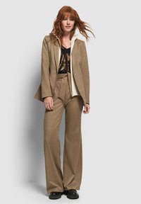 Completo su misura marrone a righe con pantaloni larghi, giacca aderente e una canotta nera sotto, abbinato a scarpe nere chunky.