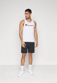 Top sin mangas de algodón blanco con el logo negro de Champion, combinado con pantalones cortos negros y zapatillas blancas. El modelo se encuentra sobre un fondo liso.