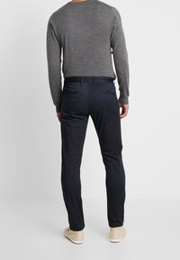 Man wearing dark slim-fit trousers, grey long-sleeve sweater, and beige casual shoes standing against a plain white background.