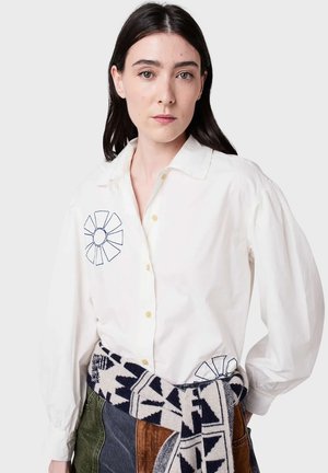 Chemise en coton blanche avec un design floral brodé sur le côté gauche de la poitrine et l'ourlet droit, présentant un devant à boutons et des manches bouffantes.