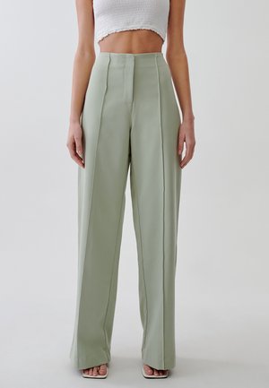 Vrouw draagt lichtgroene, hooggetailleerde, wijde broek en een wit, kort, mouwloos topje, staand voor een effen achtergrond.