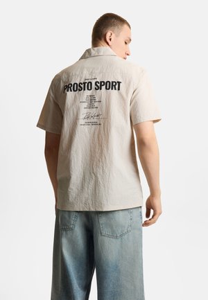 Młody mężczyzna w jasnobeżowej koszuli z krótkim rękawem z napisem „PROSTO SPORT” i jasnoniebieskich dżinsach, stojący tyłem do białego tła.