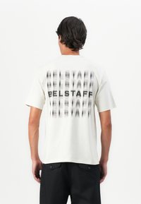 Mann iført en hvit kortermet T-skjorte med uskarp svart "BELSTAFF"-tekst gjentatt vertikalt på ryggen, kombinert med svarte bukser.