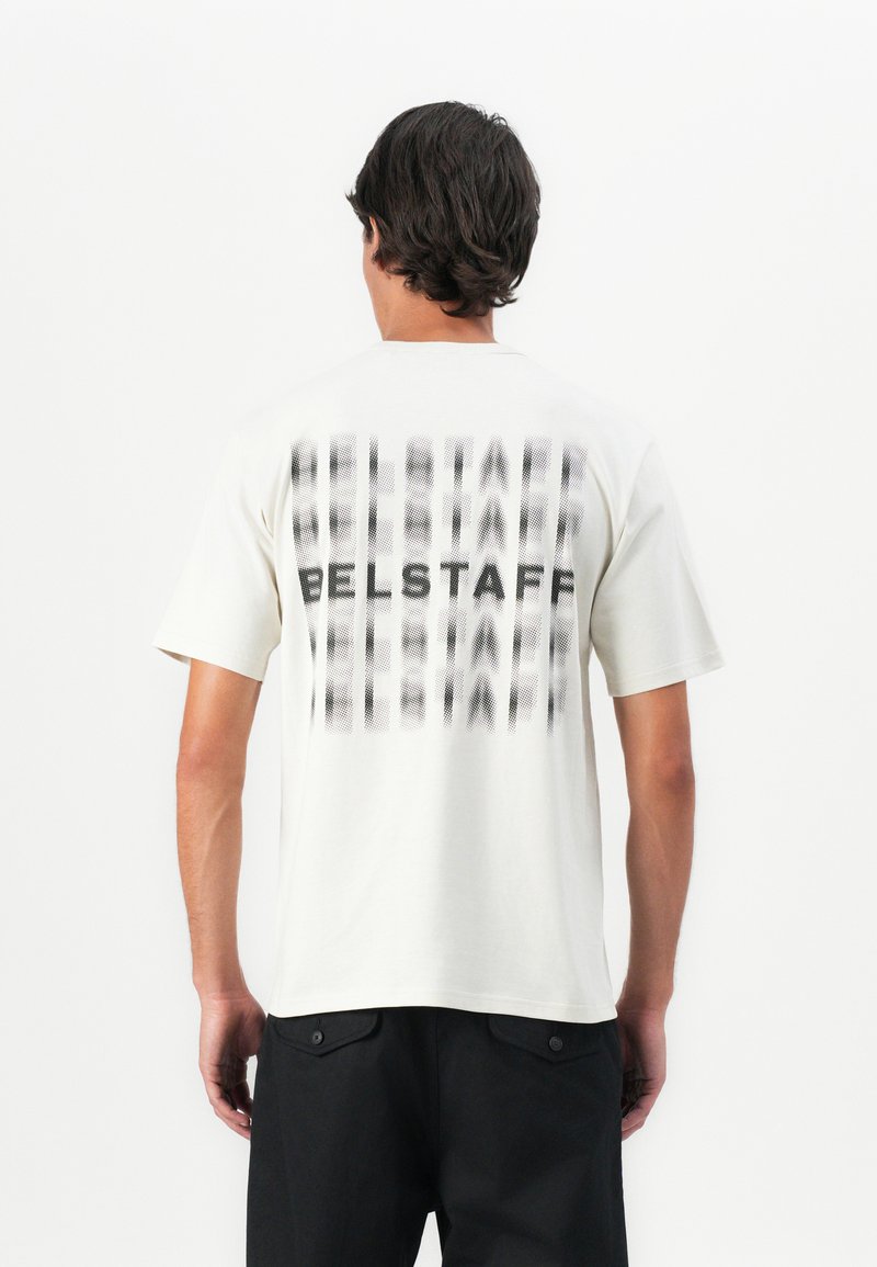 Mann iført en hvit kortermet T-skjorte med uskarp svart "BELSTAFF"-tekst gjentatt vertikalt på ryggen, kombinert med svarte bukser.