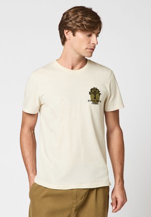 Mann trägt beige T-Shirt mit kleinem Baumgrafik und Text "SUN & TREES FAGUO" auf der Brust, kombiniert mit brauner Hose, schaut nach rechts.