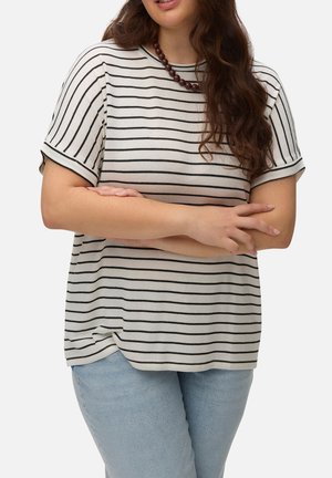 Femme aux longs cheveux ondulés portant un haut blanc et noir rayé à manches courtes, un jean bleu et un collier sombre en perles, les bras croisés.