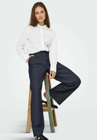 Chemise blanche à boutons avec un col agrémenté de perles, associée à un pantalon large noir, portée avec des mocassins noirs.