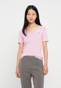 Ανοιχτό ροζ κοντομάνικο t-shirt σε σχήμα V, φτιαγμένο από μαλακό ύφασμα, με ένα μικρό κεντημένο λογότυπο στην αριστερή πλευρά του στήθους. Συνδυάζεται με γκρίζες παντελόνες.