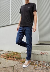 Osoba v černém tričku Calvin Klein, tmavě modrých džínech a světlých vysokých teniskách stojí venku na betonovém schodu.