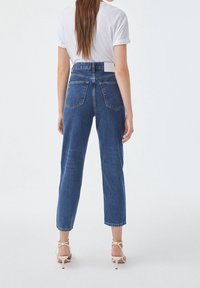 Jean en denim bleu clair avec une taille haute, coupe droite et poches arrière. Porté avec un T-shirt blanc à manches retroussées et des talons à lanières.