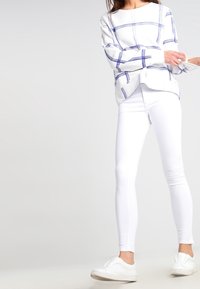 Camiseta blanca de manga larga con un patrón de cuadros azules, combinada con pantalones ajustados blancos y zapatillas blancas. La camiseta presenta un escote redondo y aberturas en los costados.