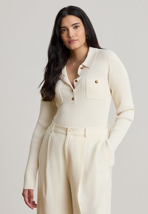 Femme aux longs cheveux foncés portant un haut côtelé crème à manches longues avec des boutons et un pantalon crème taille haute assorti, main dans la poche.