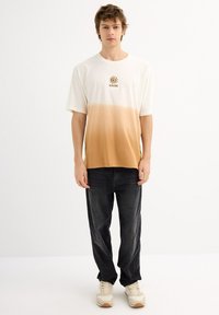 Camiseta con degradado en crema y marrón con un logo de sol. Mangas cortas, corte relajado. Combinada con jeans oscuros y zapatillas beige. Textura de tela suave.