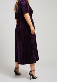 Lila sammetsmaxiklänning med korta ärmar och insydd midja, med en fladdrande fåll som når under knäet. Svarta remsandaler.