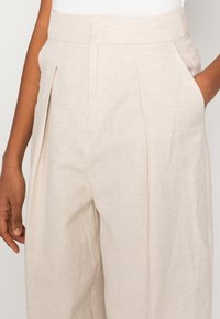 Light beige linen pants with a high waist, featuring pleats, side pockets, and a relaxed fit. The fabric is textured with a natural weave.