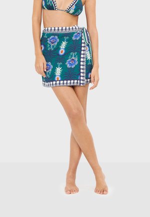 Mujer con una falda cruzada verde con estampado floral azul y blanco, combinada con un top bikini a juego, de pie descalza sobre un fondo liso.